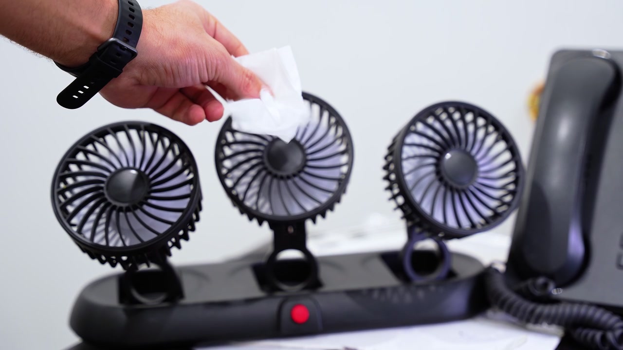 Close-up of a hand using tissue to test airflow from three compact desk fans beside an office telephone, demonstrating indoor cooling and ventilation.