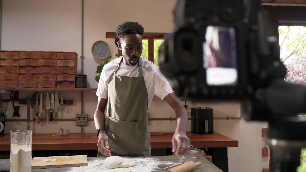 Uomo afroamericano che impasta e modella l'impasto in cucina mentre viene ripreso dalla telecamera, indossando un grembiule color oliva.