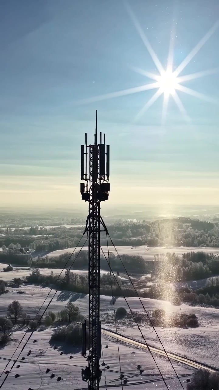 Vertical drone footage circling a telecom tower on a bright winter day, with sparkling snow crystals and a peaceful rural landscape highlighting modern communications infrastructure.