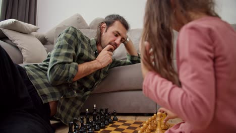 Brunette-man-in-a-green-checkered-shirt.-A-lonely-father-plays-chess-with-his-little-brunette-daughter-in-a-pink-jacket,-leaning-on-a-gray-sofa-on-the-floor-in-a-modern-apartment