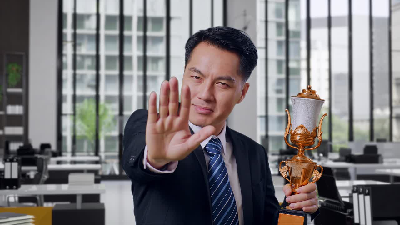 Close Up Of Asian Business Man In A Suit And Tie With A Gold Medal And Trophy Disapproving With No Hand Sign And Smiling To Camera In Office