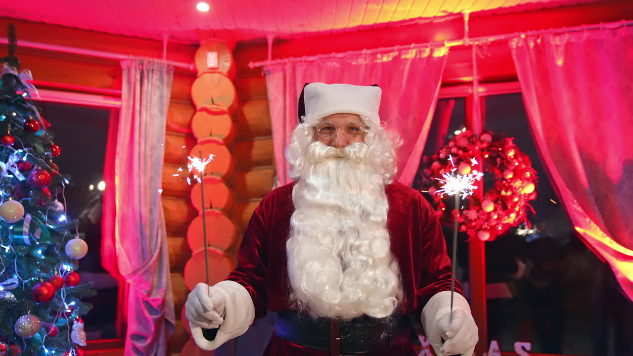 Portrait of Santa Claus indoors. Santa with white beard in glasses holding sparkling lights and looking at camera. Christmas tree with lights in decorated living room.