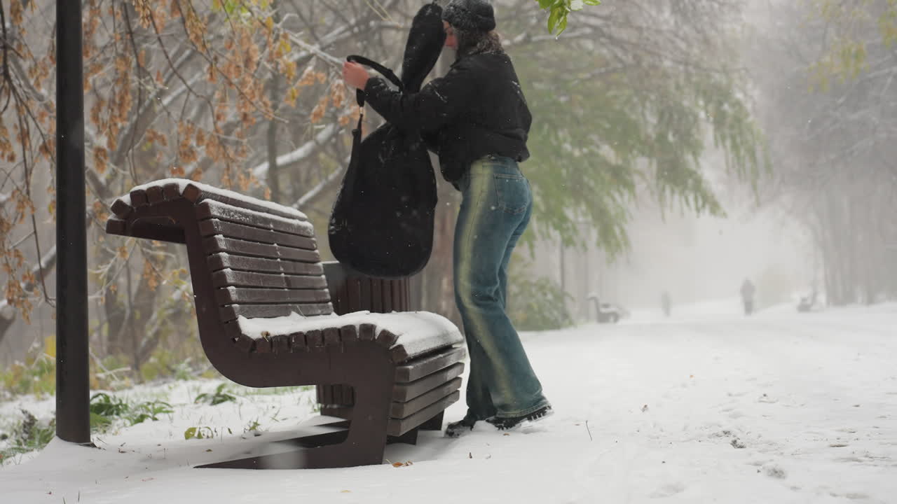 다른 한 사람 이 먼 곳 의 눈 인 배경 에서 걸어가는 동안, 평온 한 공원 에서 눈 으로 인 벤치 에 기타  을 제거 하는 여자