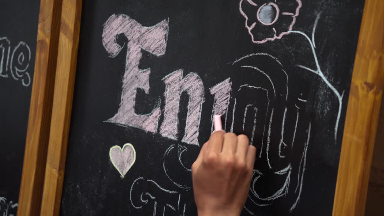 Primo piano di una mano che scrive 'goditi' con il gesso su una lavagna, aggiungendo semplici scarabocchi e scritte. Messaggio creativo, stile da menù di caffè, concetto di umore positivo e stile di vita