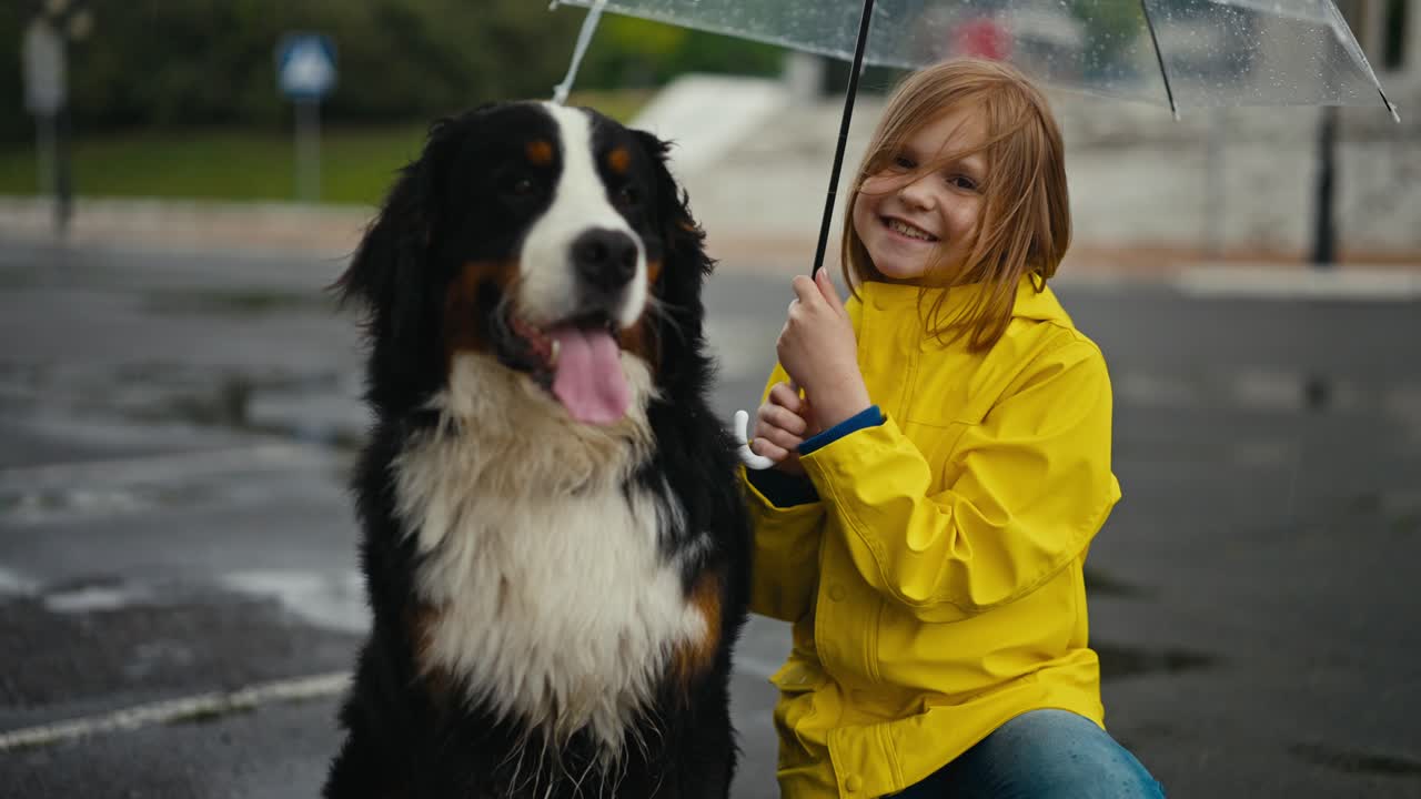Portrait of a happy blonde teenage girl in a yellow jacket and with an umbrella who poses together with her black and white rain dog in the park during