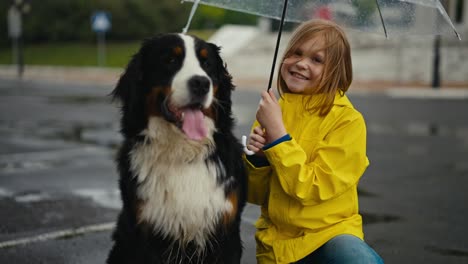 portrait of a happy blonde teenage girl in a yellow jacket and with an umbrella who poses together with her black and white rain dog in the park during