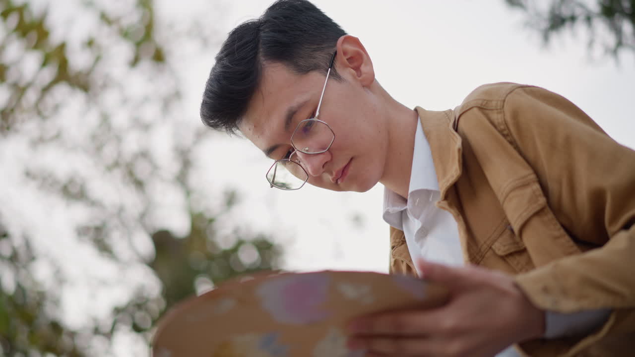 若いアジア人アーティストが屋外で絵を描いています。穏やかな都会の公園で、茶色のジャケットと眼鏡をかけ、真剣な表情でパレットと筆を持ち、紙の上で色を混ぜながらコンポジションを研究しています。