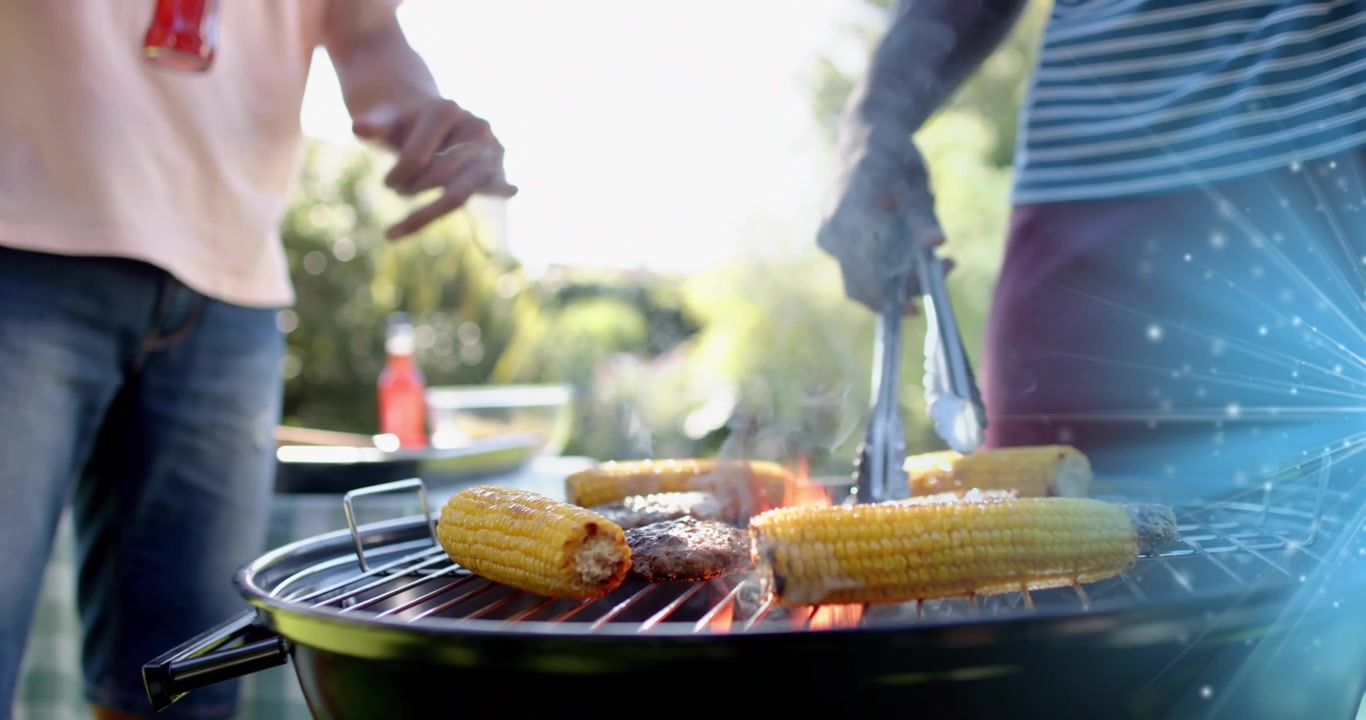 Griglia culinaria che si scalda e scintilla, uomo che usa le pinze per girare mais e hamburger mentre un amico gesticola