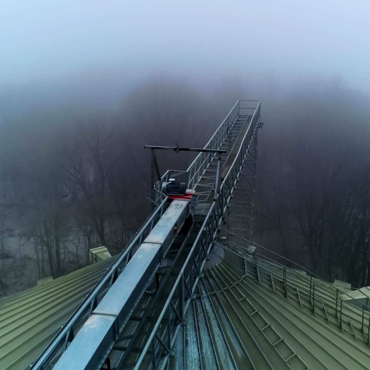Big dirty rounded top of the granary tank. Flying along the metal support connecting the elevators. Top view on the winter foggy forest