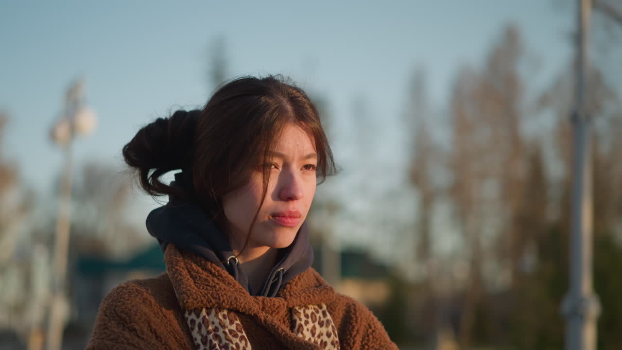 가이 아픈 소녀가 해가 지는 동안 공원을 걸어다니며, 갈색 코트와 후디를 입고, 그녀의 얼굴은 깊은 슬과 내성의 감각을 반영합니다.