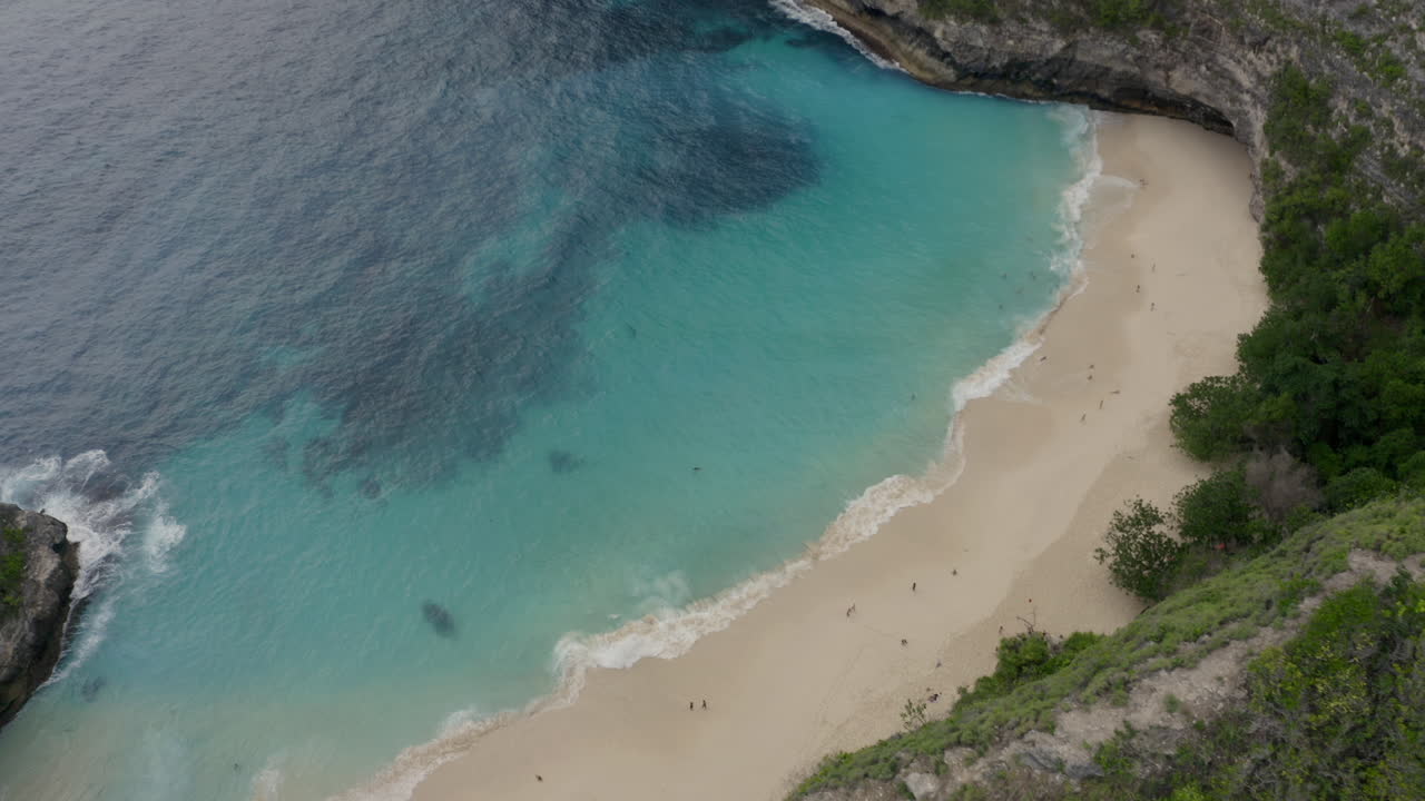 Volo sopra la cresta con un sentiero che rivela la spiaggia di Kelingking, con onde luminose che si infrangono a riva, turisti che camminano lungo la spiaggia, Nusa Penida, Bali, Indonesia. Ripresa aerea.
