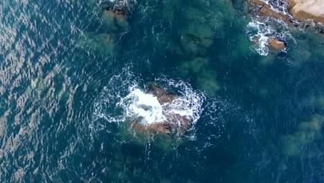 aerial shot looking directly down on the devon coastline , with waves crashing over a underwater rock and rugged coastline revealing itself