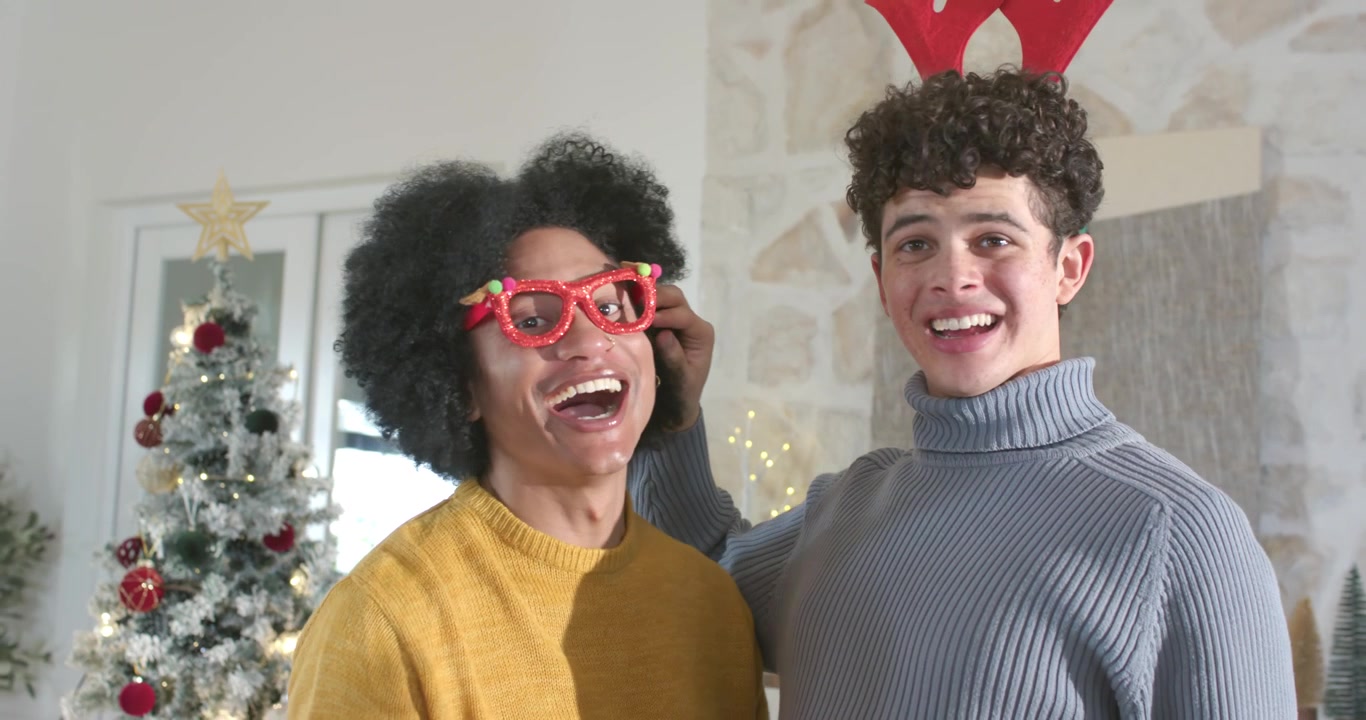 African American men in mustard, gray knits placing red glasses antlers posing at home for holidays