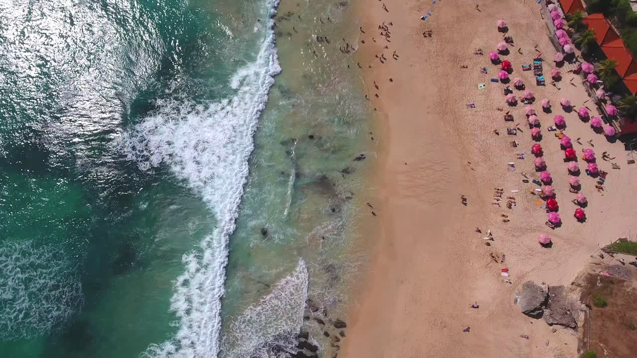 toma estática de olas rodando en la playa con sombrillas rosas, filmada en uluwatu, bali, indonesia, ambiente de vacaciones tropicales hermosa escena