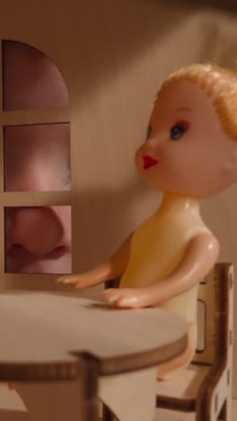 curious snide little boy stares into window of miniature handmade toy house with small dolly sitting at table near wardrobe closeup zoom out