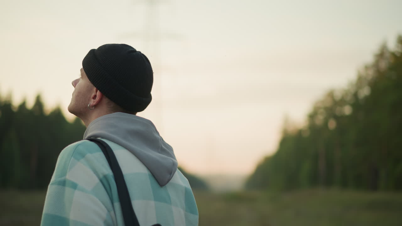 Campo lungo, escursionista caucasico con zaino che guarda l'orizzonte al tramonto, giovane pensieroso con berretto e giacca a quadri con spalline dello zaino, prato aperto e fila di pini, tenue bagliore crepuscolare, stato d'animo contemplativo di scoperta