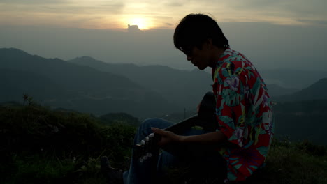 Asiatischer-Musiker-Spielt-Gitarre-Mit-Sonnenuntergangshintergrund-In-Der-Berglandschaft,-Vintage-Sonnenbrille-Und-Buntem-Hemd,-Musikalische-Inspiration