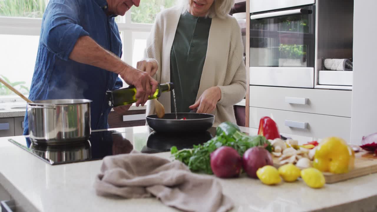 Senior man reaching and pouring olive oil into pan as woman stirring kitchen vegetables meal prep