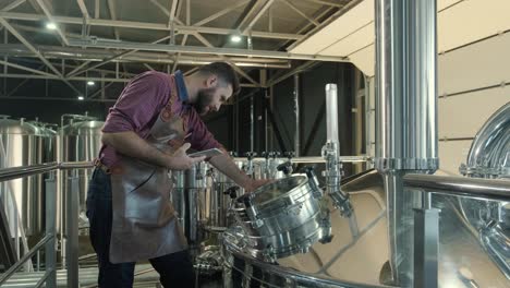 young male brewer wearing a leather apron supervise the process of beer fermentation at a modern brewery factory
