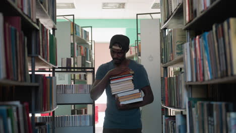 Student in casual clothing and cap walking between library shelves holding large stack of books, surrounded by rows of colorful books under bright indoor lighting