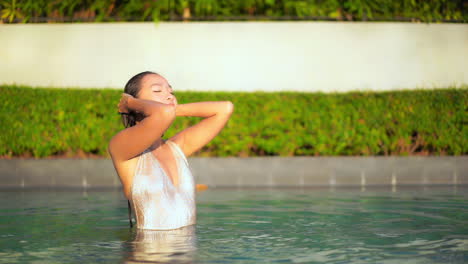 Toma-En-Cámara-Lenta-De-Una-Joven-Atractiva-Parada-Medio-Sumergida-En-La-Piscina-Tocando-Su-Largo-Cabello-Mojado-Con-Los-Ojos-Cerrados-Y-Luego-Abriéndolos-Lentamente-Con-Deleite-Y-Sonrisa-En-Su-Rostro