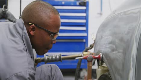 african american male car mechanic holding a screwdriver and using a hammer to repair a car