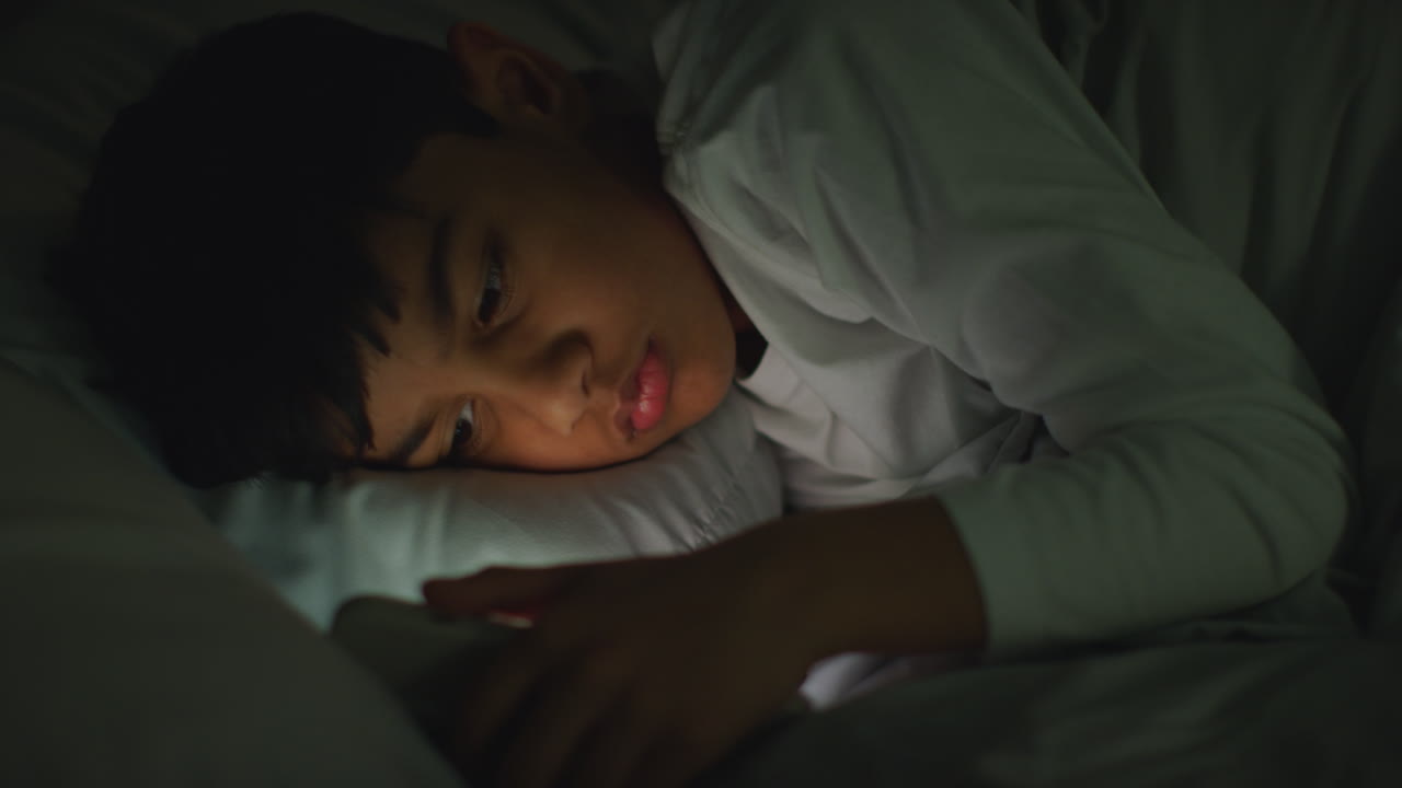 집에서 침실에 누워있는 어린 소년은 밤 4시에 문자 메시지를 보내기 위해 휴대 전화를 사용합니다.