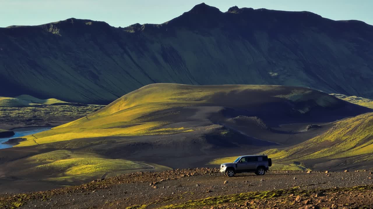 Ripresa cinematografica con drone di un SUV solitario su una cresta rocciosa negli altopiani islandesi, con colline vulcaniche illuminate dal sole e una parete montuosa scura, che evoca avventura remota e solitudine
