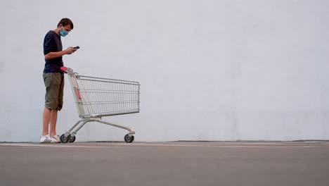 man in mask with smartphone pushing shopping cart. customer with shopping trolley along the wall of the store. guy in a disposable facemask following the dinstance immersed himself in internet surfing