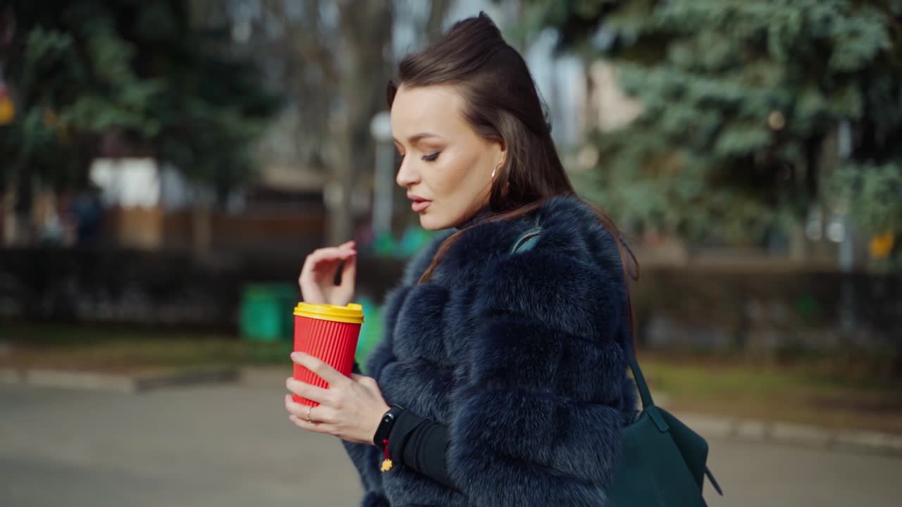 Side view of a beautiful woman outdoors. Pretty girl in luxury fur coat walking in the park with a plastic cup of coffee. Slow motion.