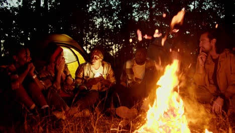 evening cozy communication near the fire, a small group sits near the fire and tells scary stories against the backdrop of the floor and the evening autumn forest. a man in a hat shines a flashlight in his face and tells scary stories to a group during a hike