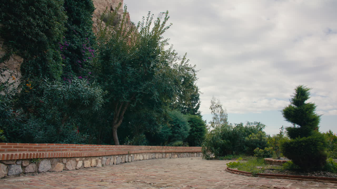 Wide shot of a stone walkway bordered by dense greenery and a brick wall, under a calm, overcast sky