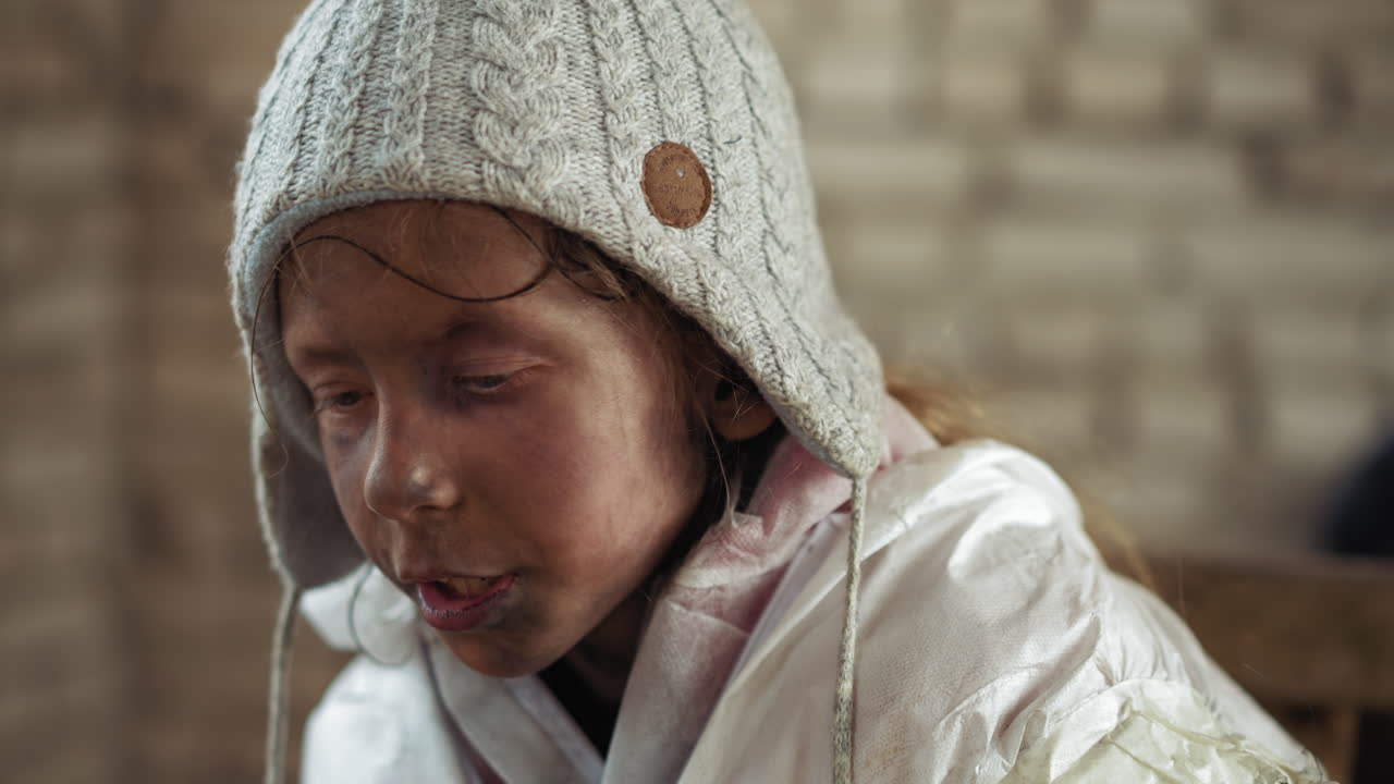 Dirty child dressed in thick white jacket and warm knitted hat with serious facial expression engages in conversation, showing signs of hardship and resilience in cold place