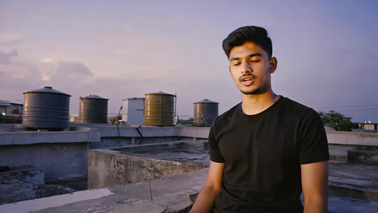 Young man practicing meditation on a rooftop at sunset, with water tanks in the background, promoting mindfulness and inner peace in urban environment