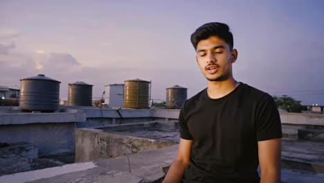 young man practicing meditation on a rooftop at sunset, with water tanks in the background, promoting mindfulness and inner peace in urban environment