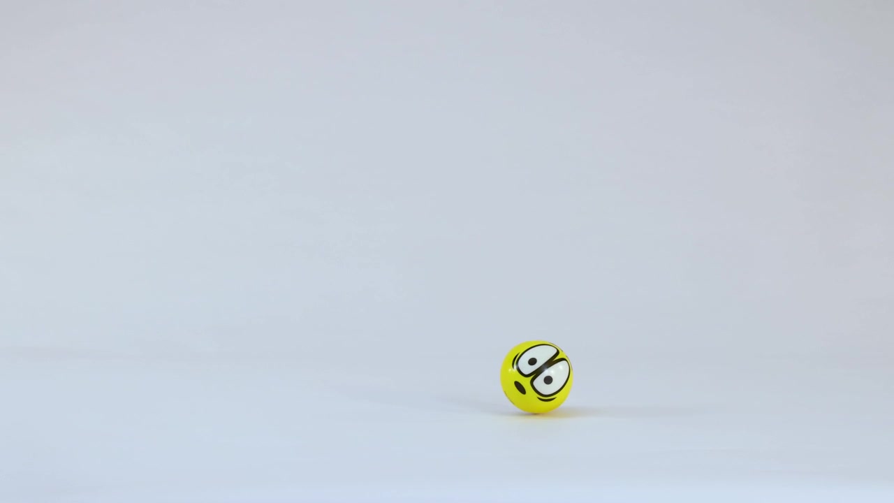 Una palla gialla con un'emoji rimbalza e rotola sul pavimento bianco di uno studio