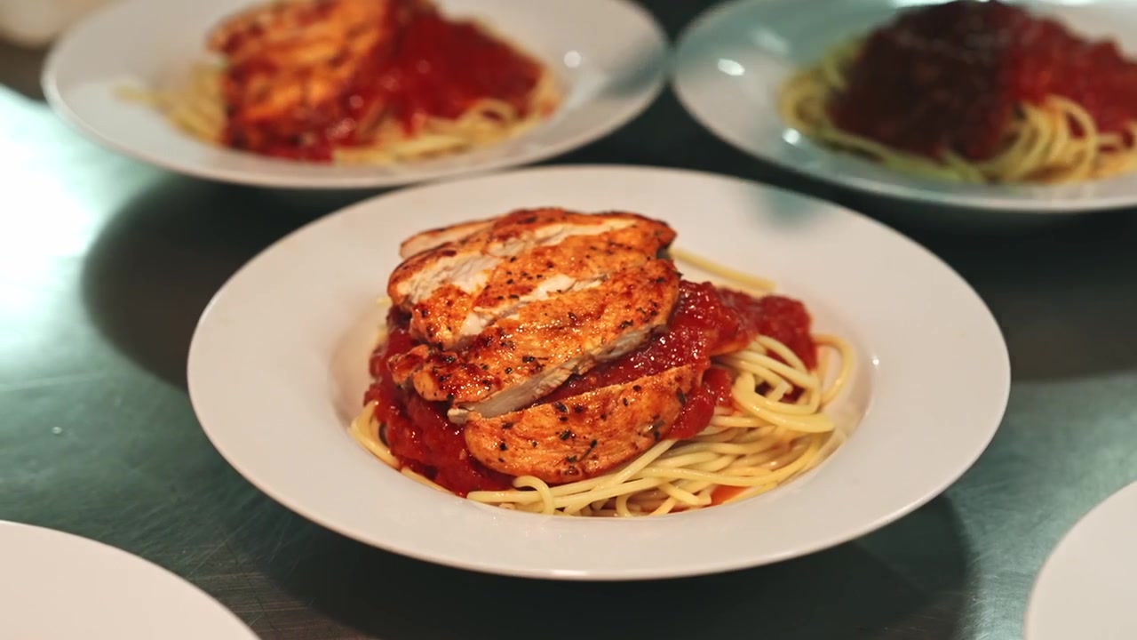 Ripresa ravvicinata in movimento di uno chef che indossa guanti neri mentre affetta pollo grigliato condito su un tagliere e posiziona il pollo affettato sopra piatti di spaghetti al pomodoro