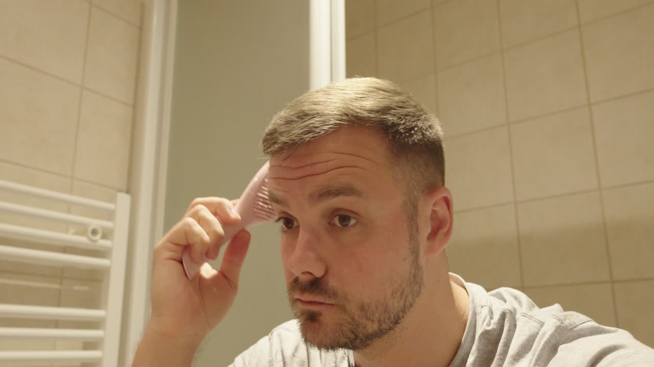 A young man styles his short haircut using a pink brush in front of the bathroom mirror. Morning grooming routine in a tiled, modern bathroom setting