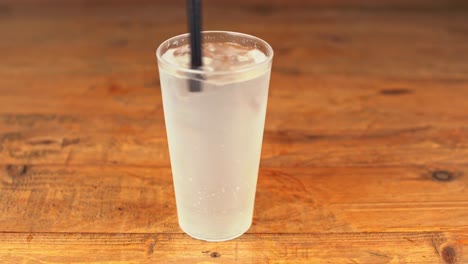 Close-up-of-a-refreshing-glass-of-mineral-water-with-ice,-a-straw,-and-a-touch-of-lemon-being-stirred-on-a-wooden-table-in-a-bar