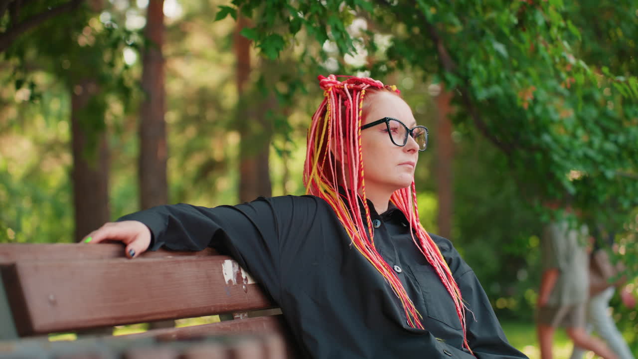 profesora caucásica con trenzas brillantes reclinada en un banco del parque, postura relajada con el brazo a lo largo del respaldo, observando árboles iluminados por el sol, pausa reflexiva entre clases, conjunto negro informal y gafas