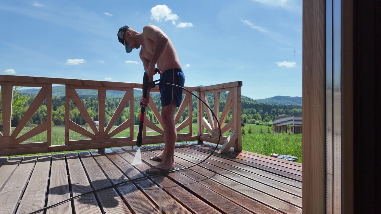 Operaio a torso nudo in pantaloncini blu pulisce una terrazza in legno con un'idropulitrice, con erba, foresta e cime montuose visibili sullo sfondo.