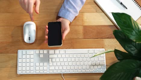 Hand-of-businesswoman-using-mobile-phone-at-desk-with-keyboard-and-mouse-on-table