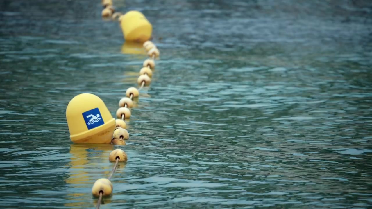 Yellow buoys with a swimming icon floating on water, marking a safe beach swimming area. Ripples visible on water surface