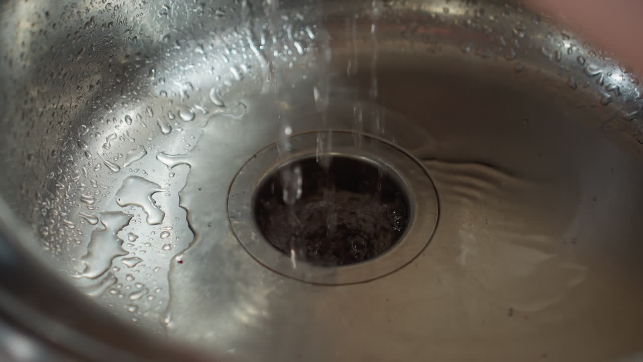 Water droplets drip into stainless steel sink creating ripples and reflections on shiny metal surface, macro view emphasizes water flow and texture with subtle reflections