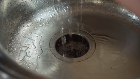 water droplets drip into stainless steel sink creating ripples and reflections on shiny metal surface, macro view emphasizes water flow and texture with subtle reflections