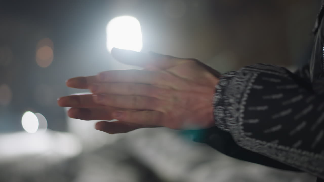 Close up of individual rubbing hands together to stay warm on cold night, soft light glare and bokeh in background, fingers gently pressing with tension and chill reflected in motion and tone
