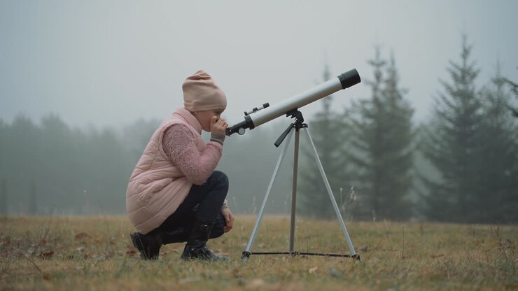 Barn interesseret i astronomi og videnskab. Lille pige kigger gennem et teleskop i naturen, iført vintertøj