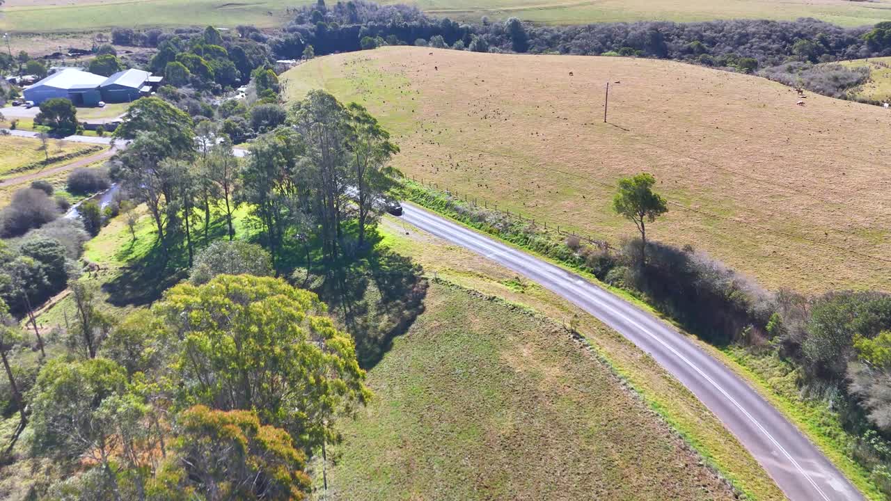 Drone footage follows a single car traveling along a winding road through grassy fields and scattered trees under bright daylight in rural Australia
