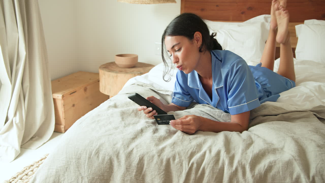 Smiling woman in pajamas shopping online with credit card on bed at home