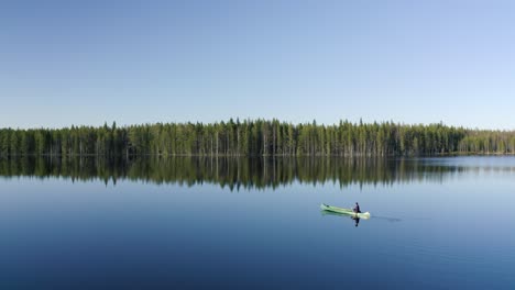 Mann-Im-Grünen-Kajak-Paddelt-An-Einem-Sonnigen-Tag-Ruhig-über-Einen-Sehr-Glatten-See,-Der-Den-Waldrand-Inmitten-Der-Schönen-Natur-Finnlands-Widerspiegelt